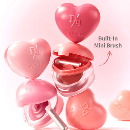 Heart-shaped dasique lip balm with a built-in mini brush on a pink background